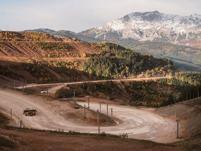 Vue d'une route sinueuse en terre dans un paysage de montagne avec un véhicule jaune et une montagne enneigée en arrière-plan.