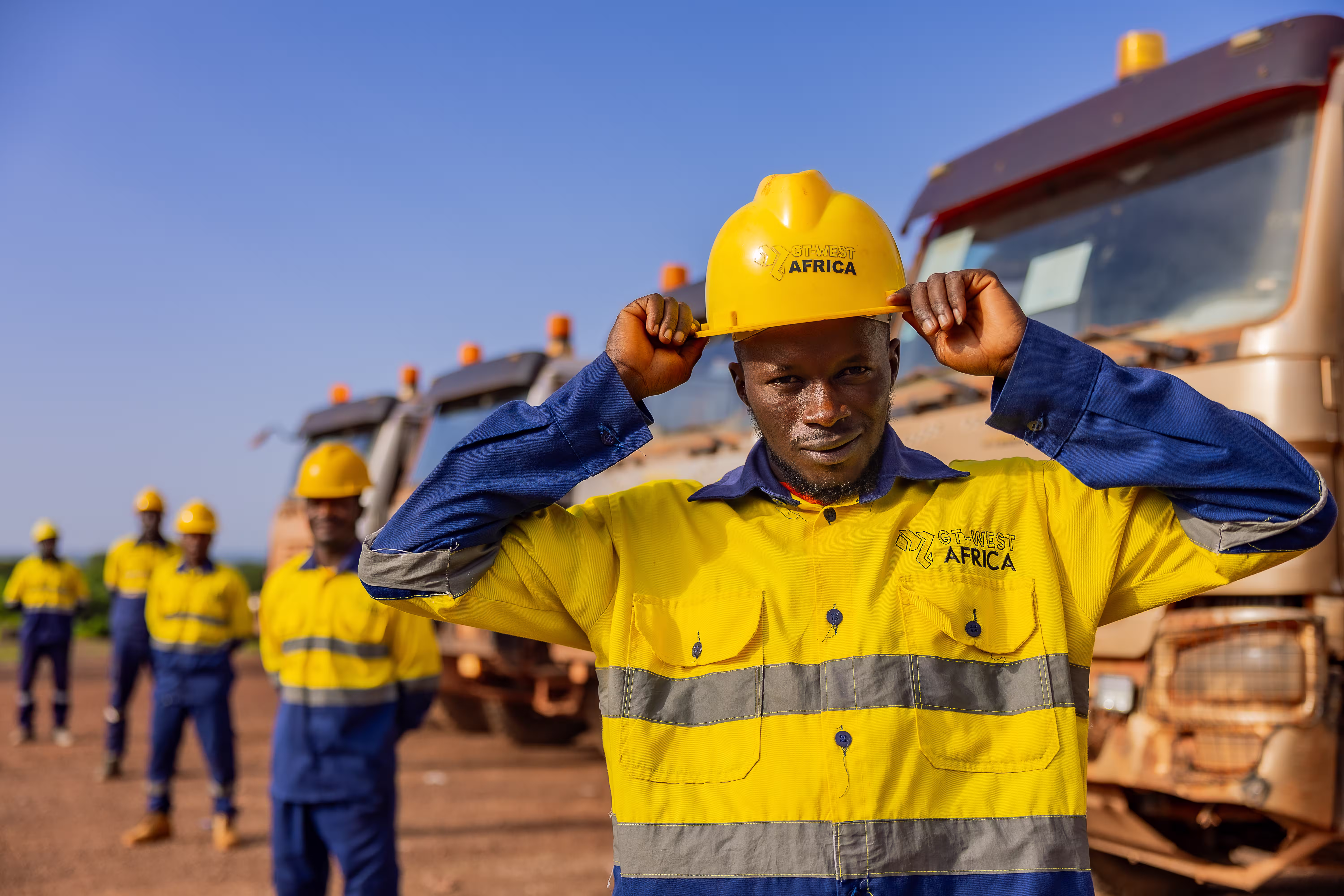 Travailleur portant un casque jaune GT-WEST AFRICA devant des camions avec des collègues en arrière-plan.