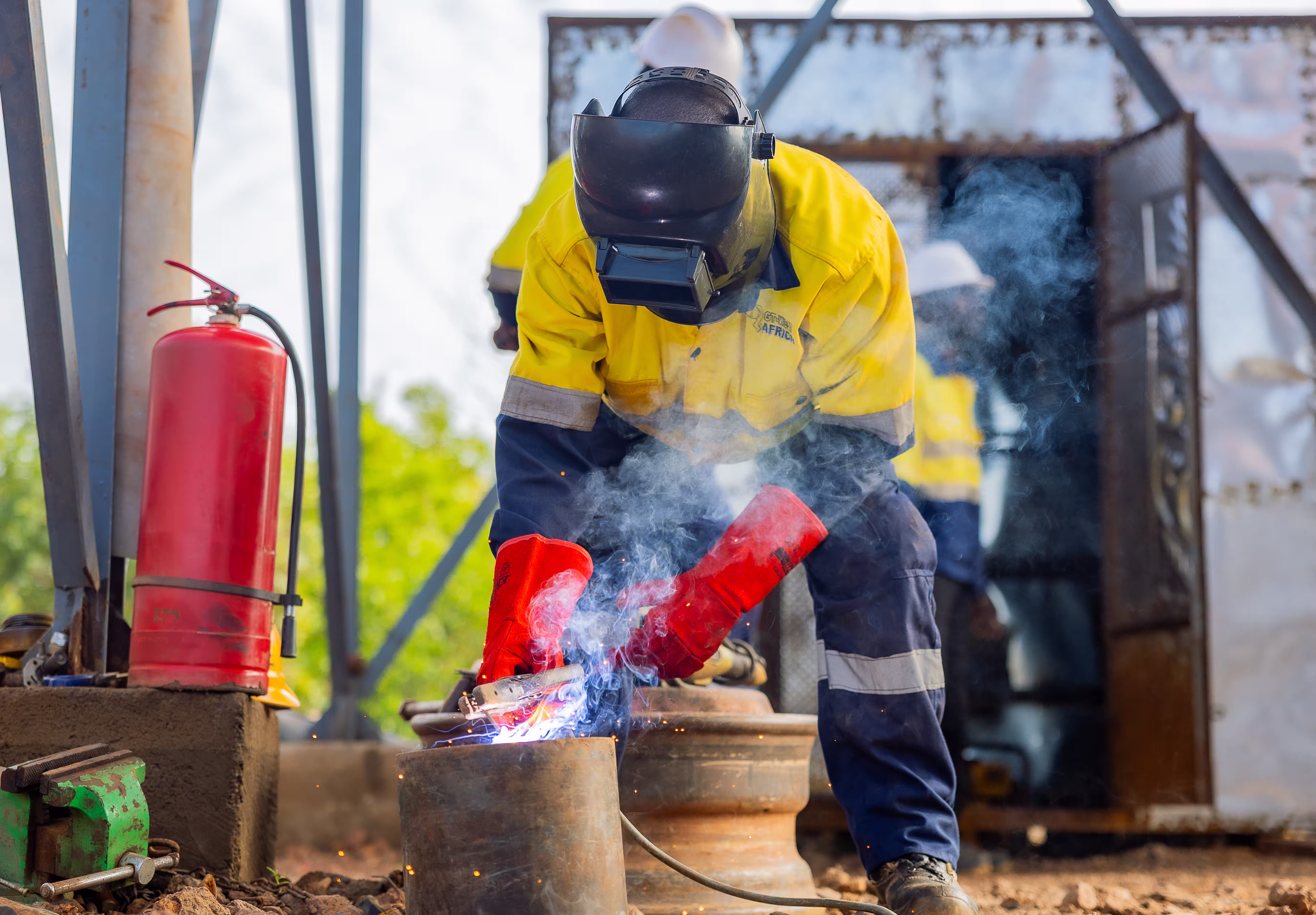 Ouvrier soudant du métal avec équipement de protection jaune et gants rouges dans un atelier extérieur.