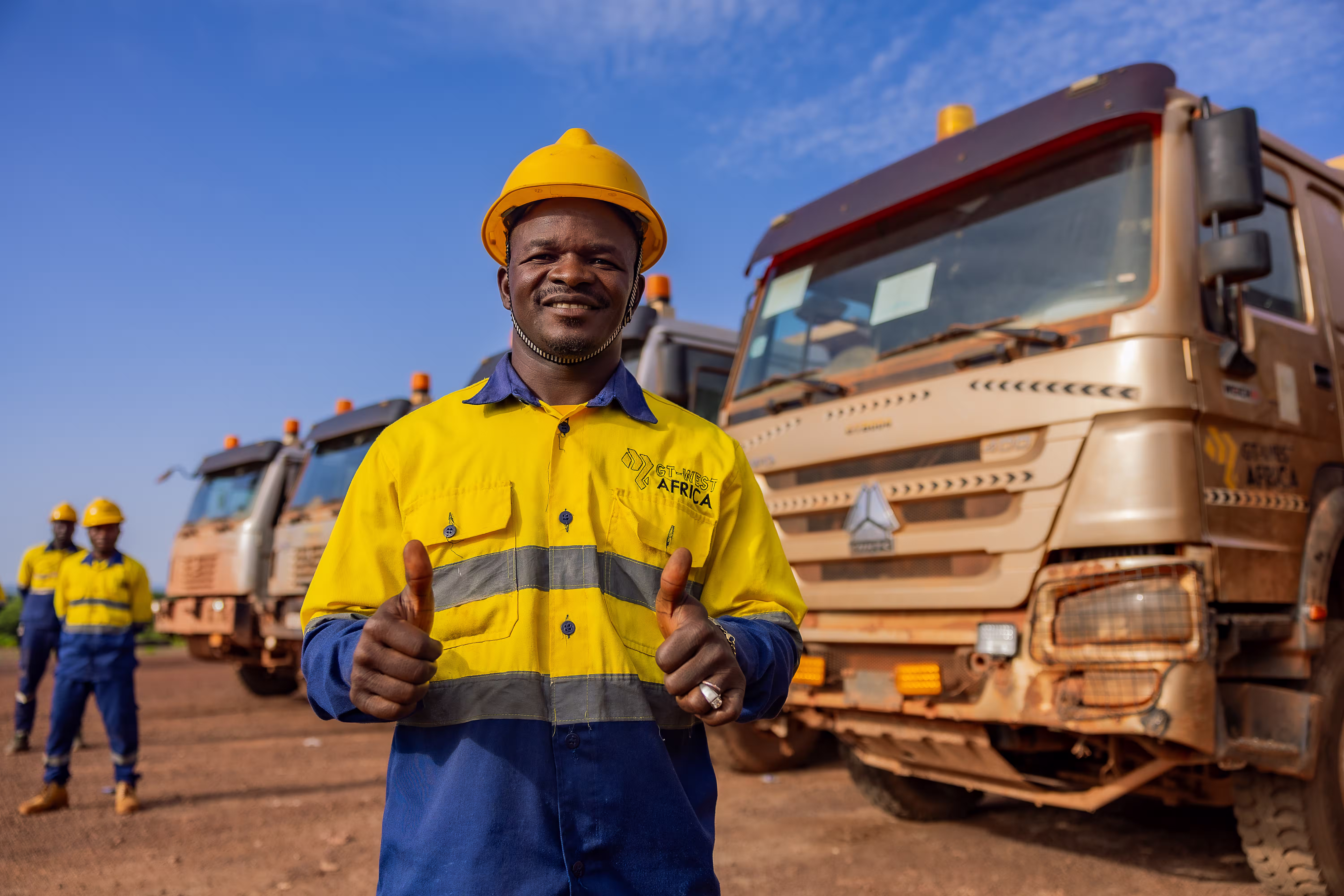 Ouvrier en casque jaune et uniforme jaune et bleu fait un signe de pouce en l'air devant des camions.