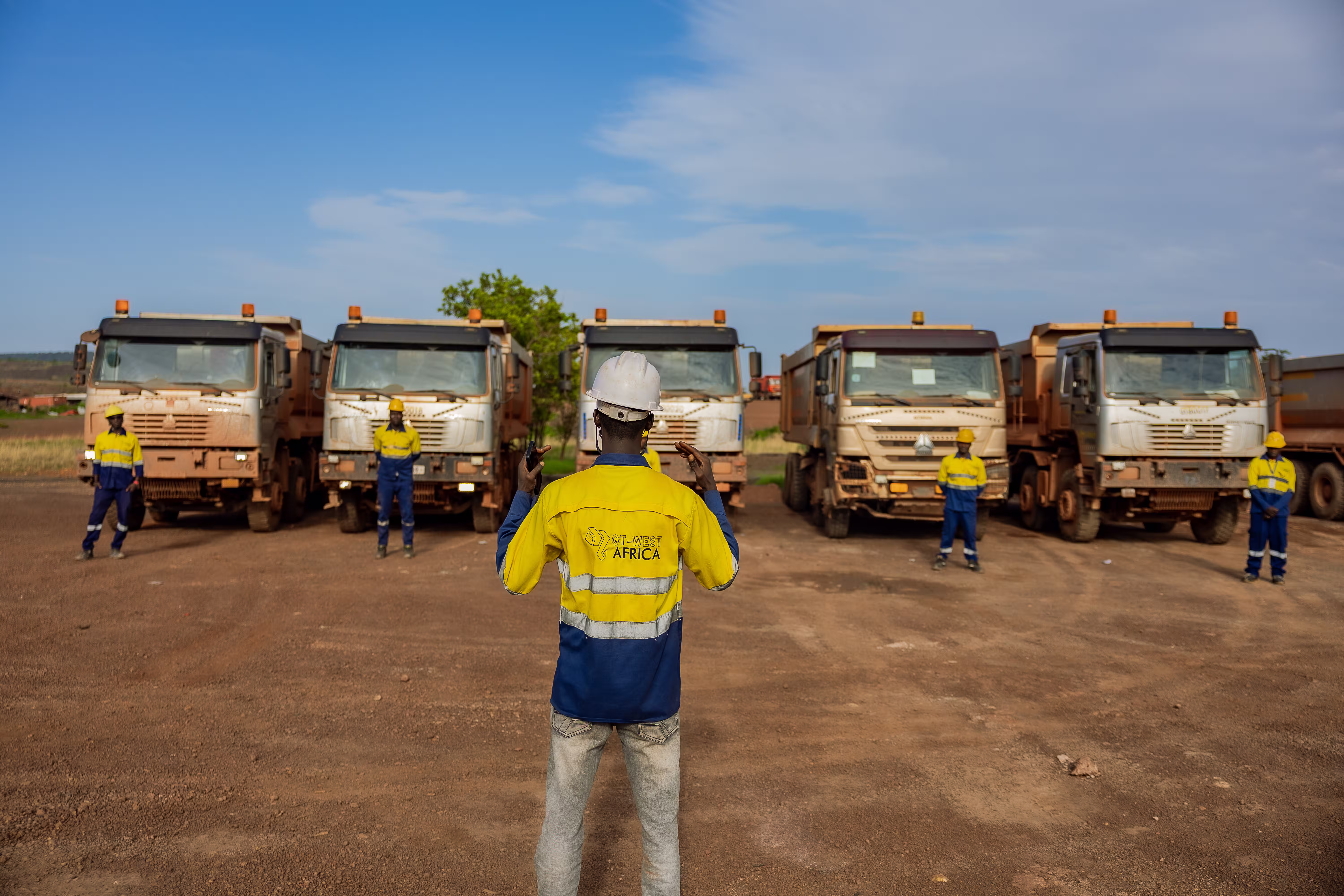 Un homme en tenue de sécurité fait face à cinq camions et quatre ouvriers en casque sur terrain poussiéreux.