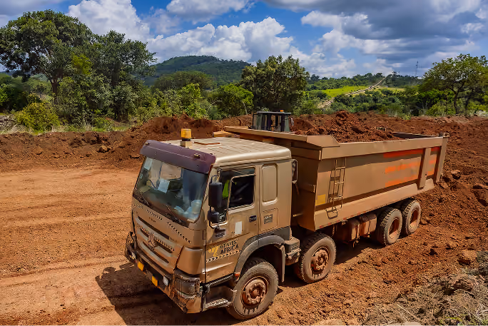Un camion-benne beige chargé de terre sur un chantier entouré d'arbres et de collines.