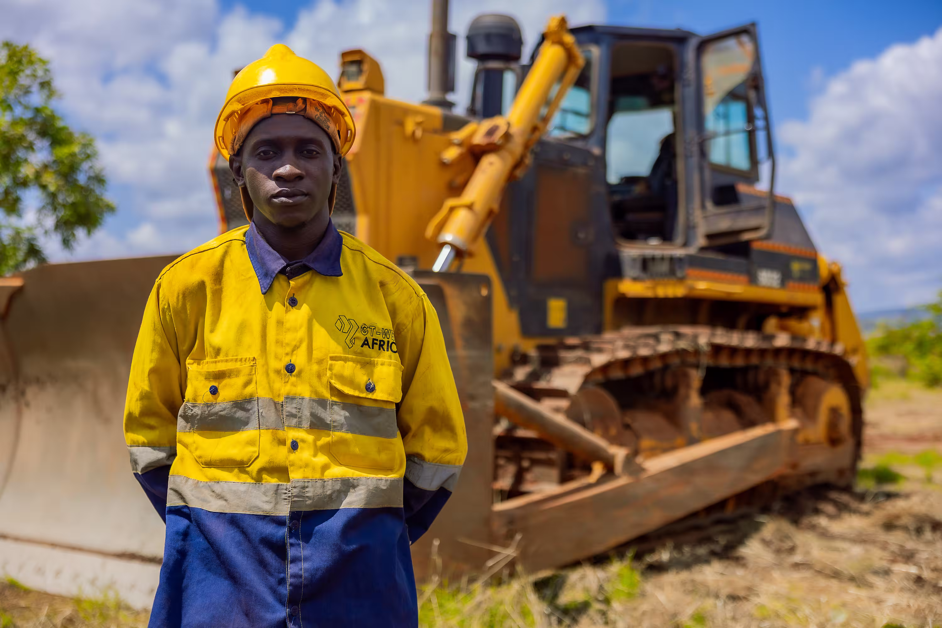 Ouvrier en casque et tenue jaune-bleue debout devant un bulldozer en plein air.