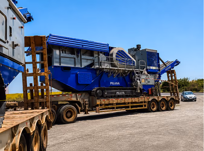Machinerie industrielle bleue marquée Pilana chargée sur une remorque sur une route ensoleillée.