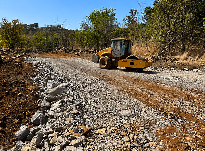 Engin jaune compacteur sur une route en construction avec des pierres et arbres autour.