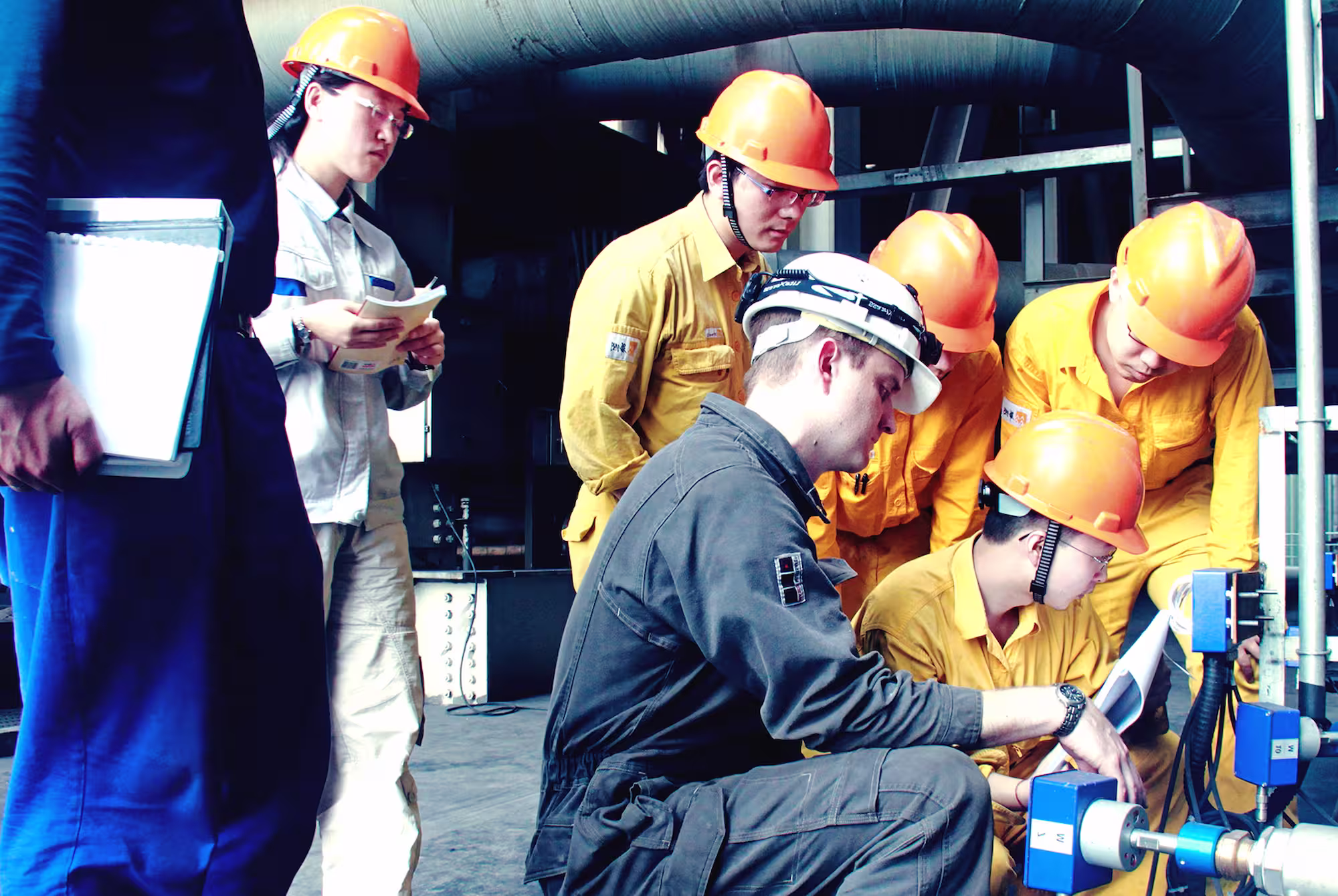 Group of engineers in safety helmets inspecting industrial pipe equipment during a worksite briefing.