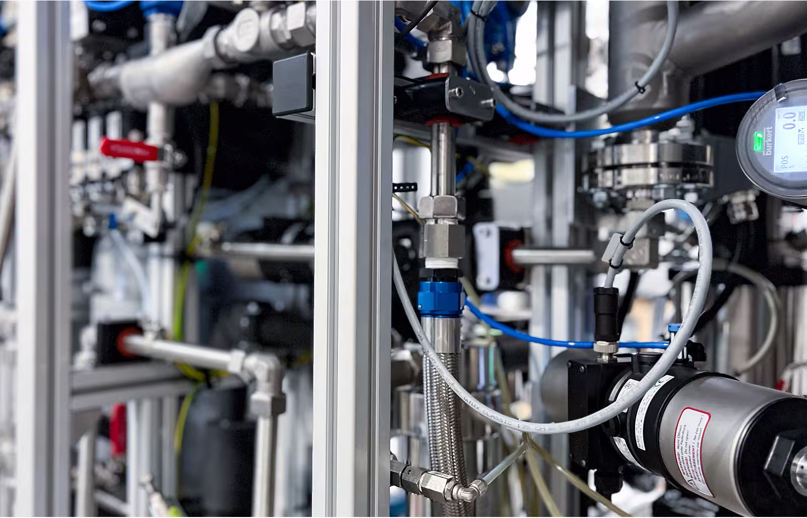 Close-up of industrial equipment with metal pipes, connectors, valves, and blue cables in a factory setting.
