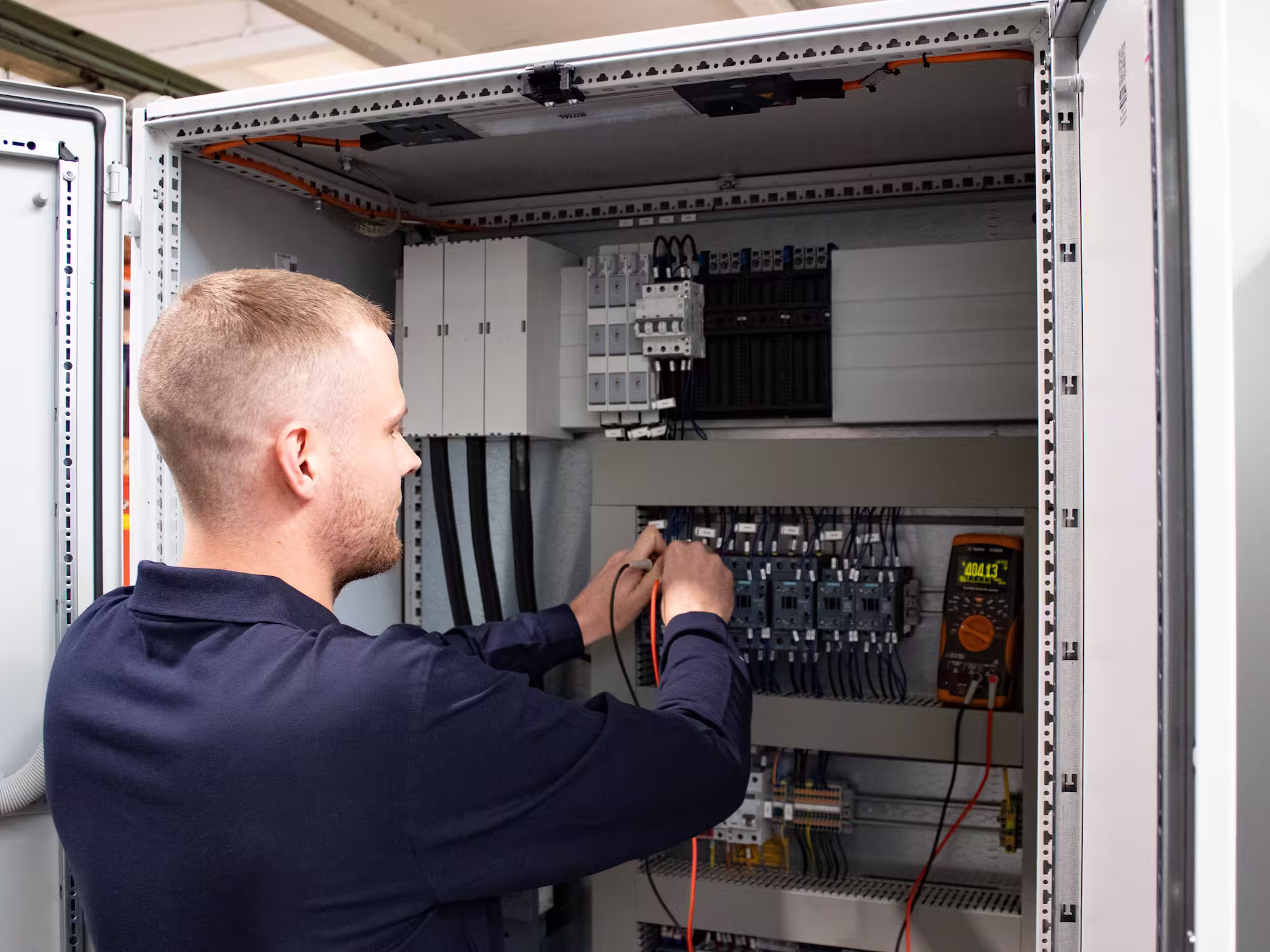 Technician testing electrical wiring inside an open control panel with digital multimeter.