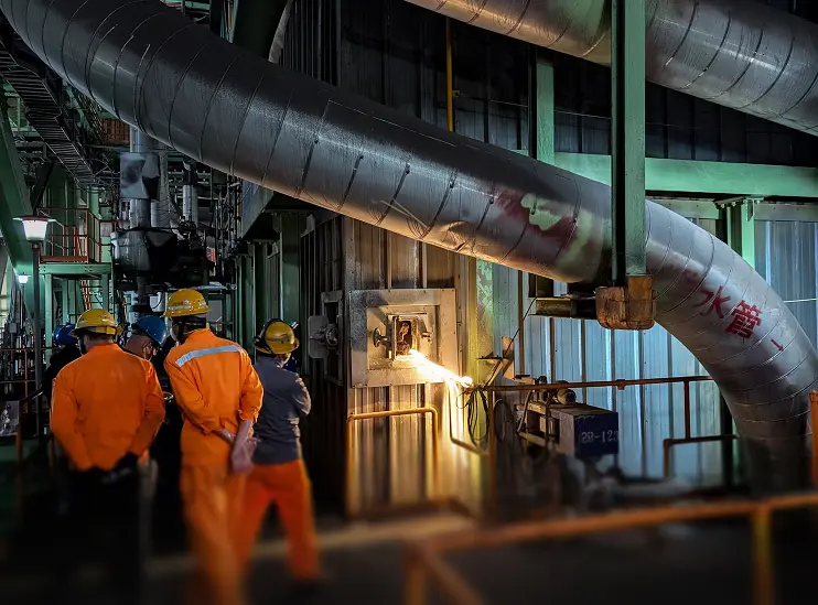 Workers in orange safety gear and helmets observe a large industrial furnace with glowing fire inside.