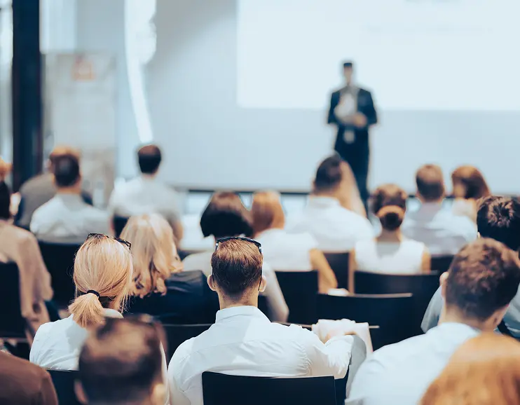 Audience seated facing a speaker giving a presentation in a conference or seminar room.