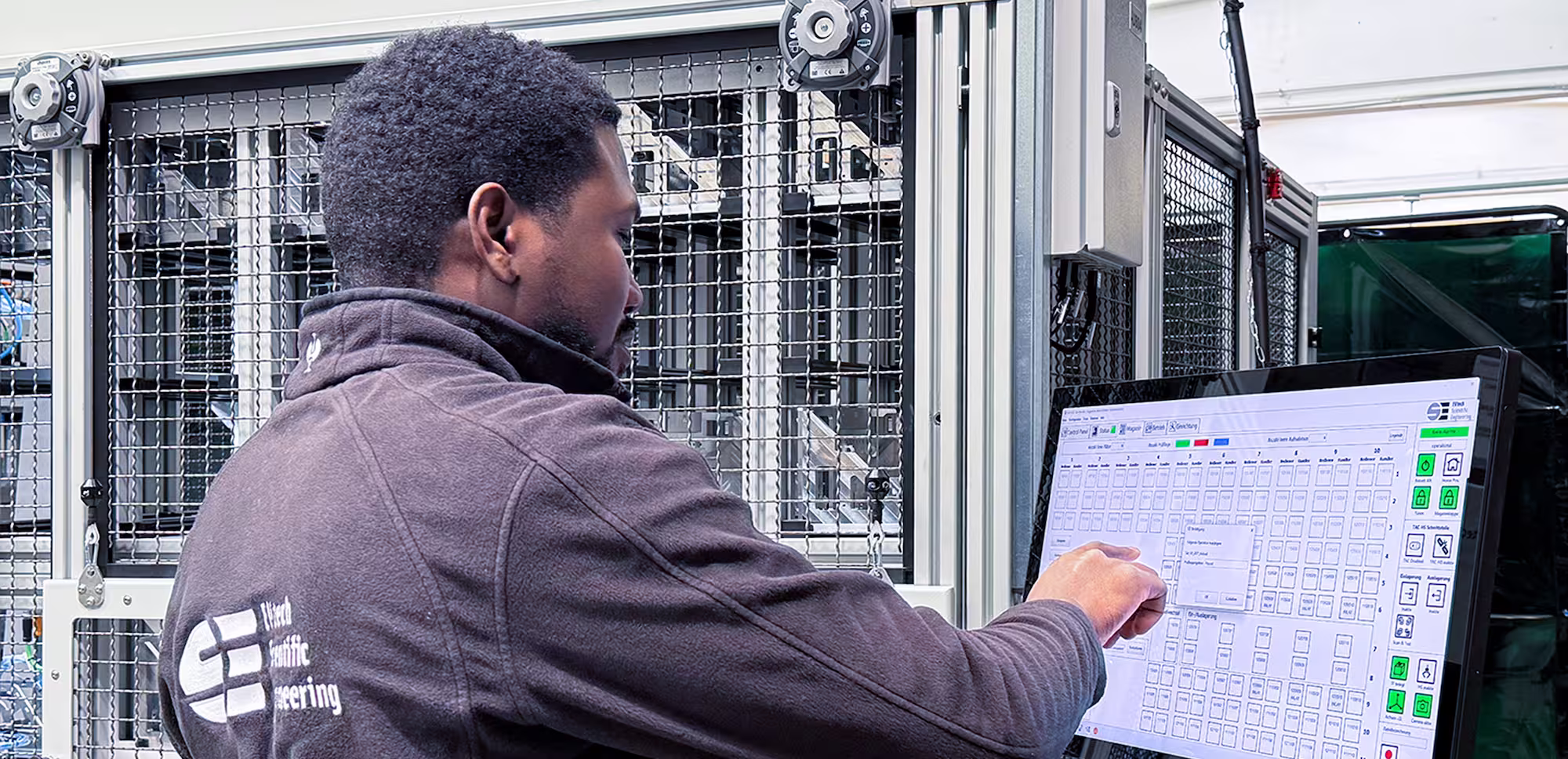 Man operating a touch screen control panel in an industrial setting with machinery behind him.