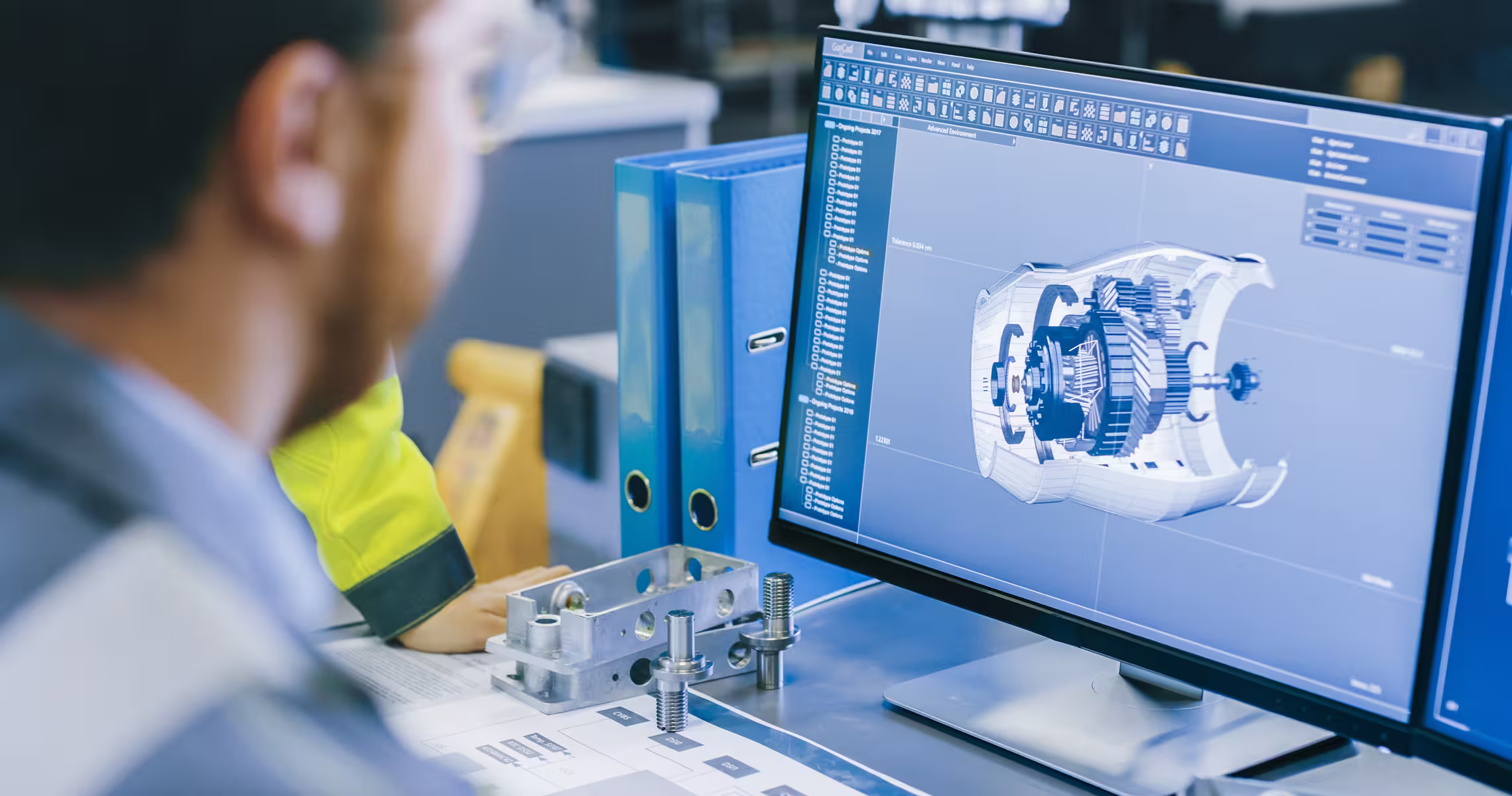 Two engineers examining a mechanical part model displayed on a computer screen in an industrial workspace.