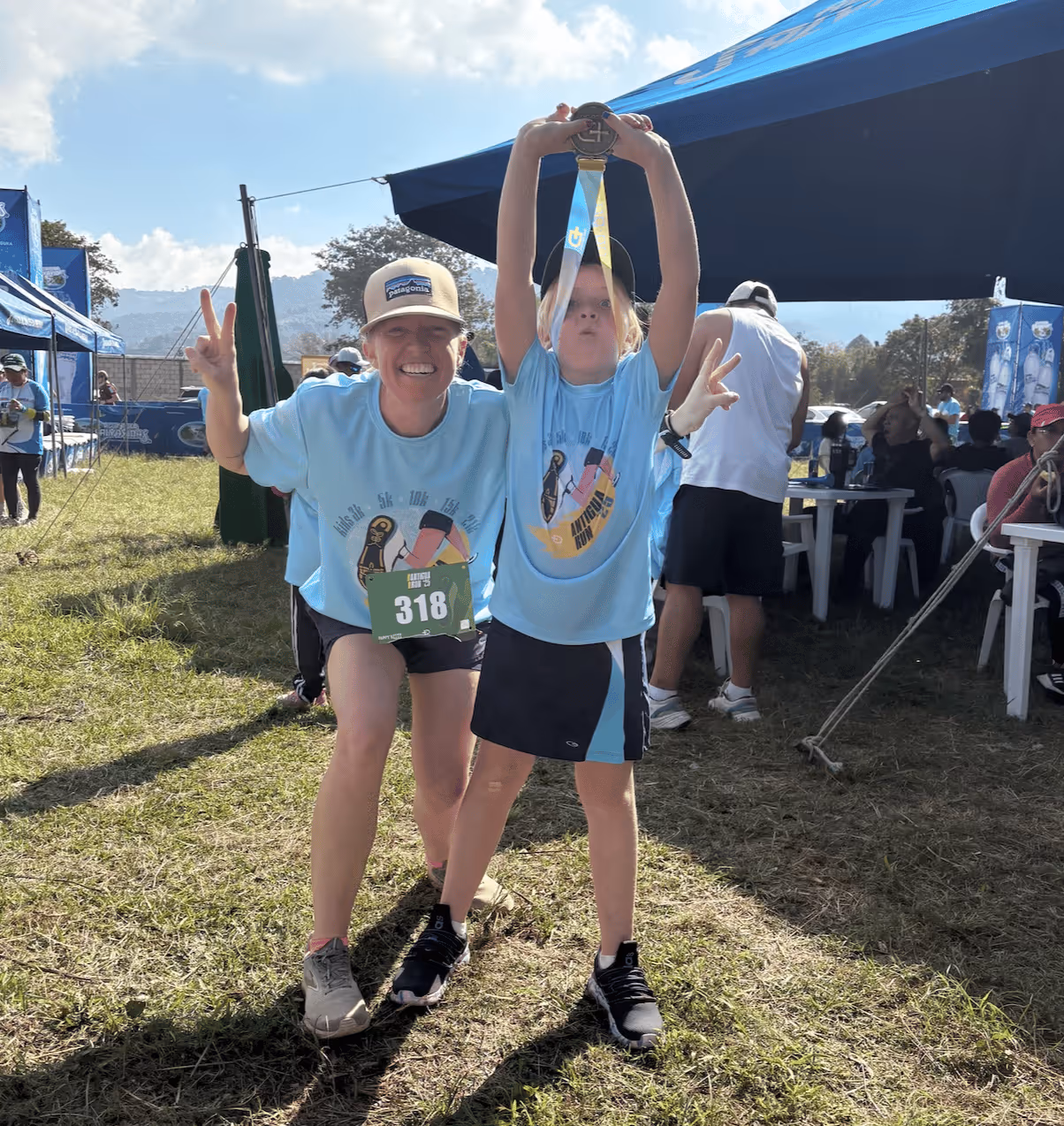 Smiling woman and child in race shirts hold medal and show peace signs at outdoor event tent.