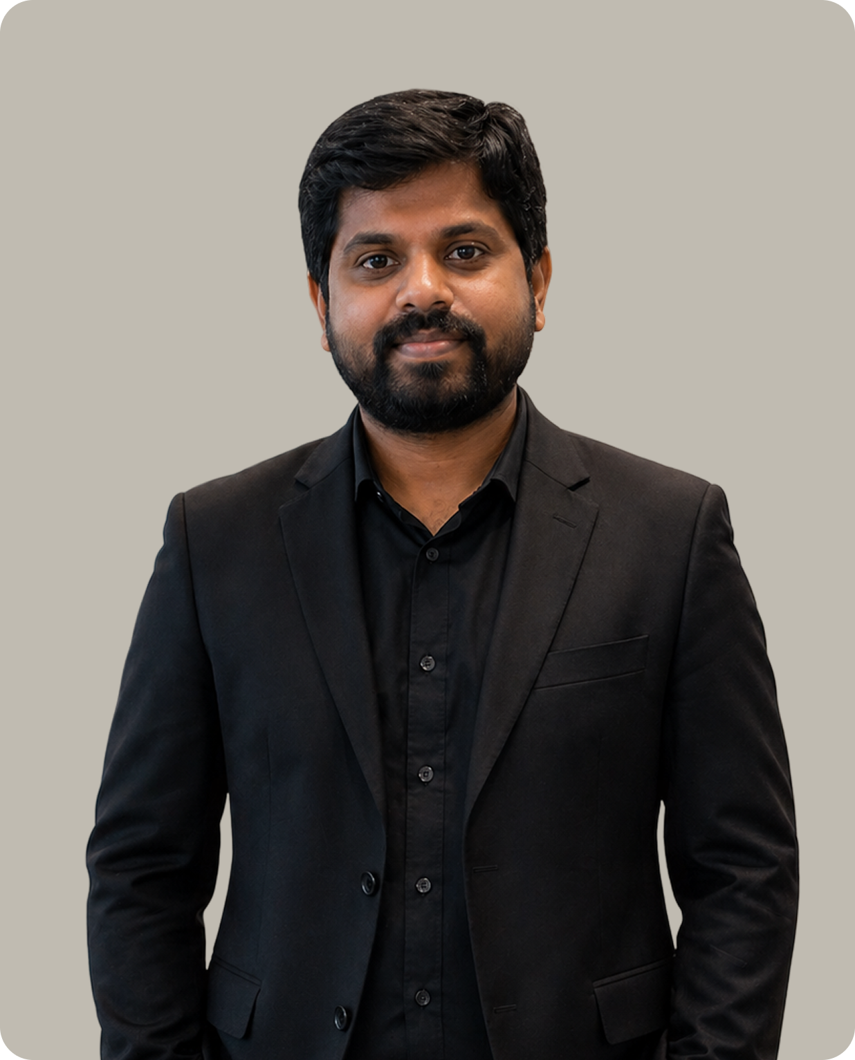 Smiling man with dark hair and beard wearing a black suit and black shirt against a plain background.