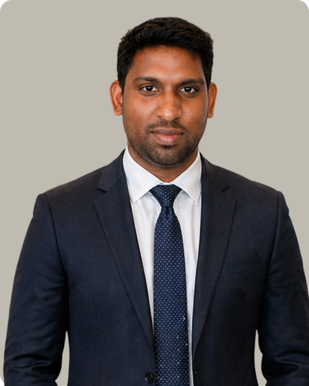 Man wearing a dark navy suit, white shirt, and dark blue dotted tie standing against a plain light gray background.