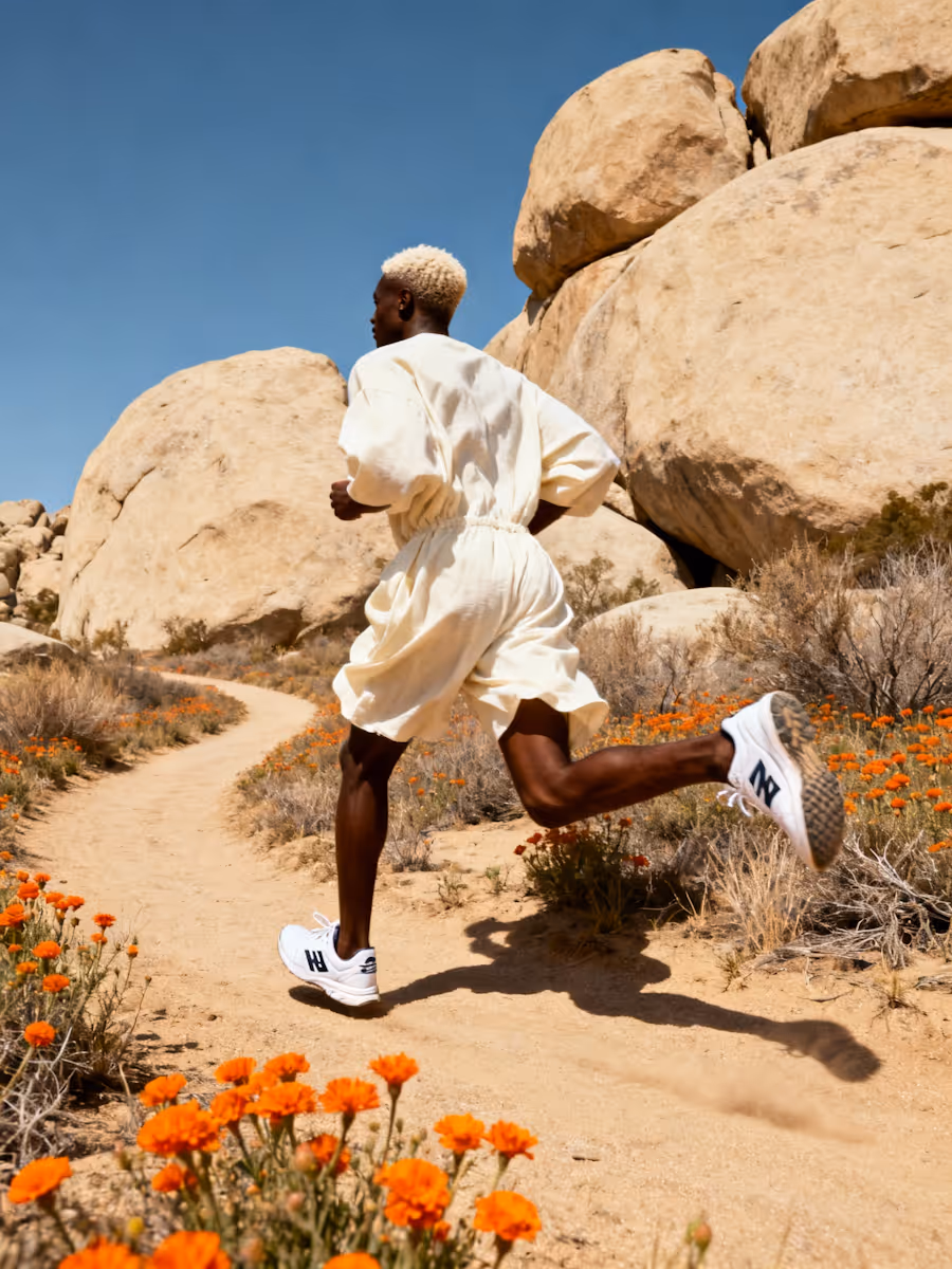 Person with blonde hair wearing white running shoes and light clothing jogging on a desert trail surrounded by orange flowers and large boulders.