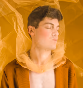 Young man with closed eyes wearing a brown jacket and wrapped in translucent orange fabric.