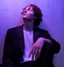 Person with curly hair wearing a black blazer and white shirt, looking up against a textured wall with purple lighting.