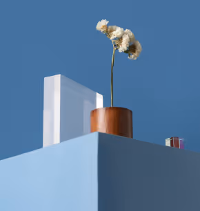 White flowers in a small wooden vase on a blue surface with a clear acrylic block and a small colorful object against a blue background.