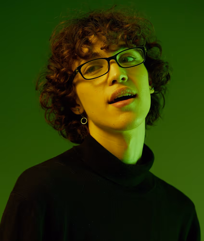 Person with curly hair wearing glasses and a black turtleneck against a green background.