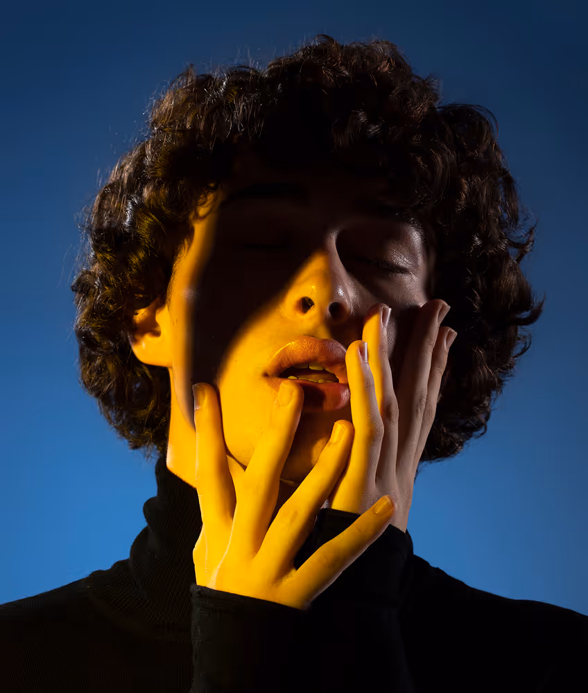 Person with curly hair wearing a black turtleneck, eyes closed, hands touching face, yellow light illuminating parts of their face and hands against a blue background.