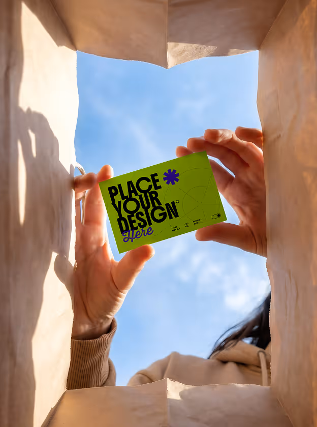 Person holding a green card with the text 'Place Your Design Here' against a blue sky, viewed from inside a paper bag.