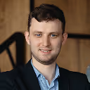 Portrait of a young man with short dark hair and a trimmed beard, wearing a blue shirt and dark jacket, against a wooden background.