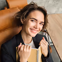 Smiling woman with short brown hair sitting on a couch holding a notebook and pen.