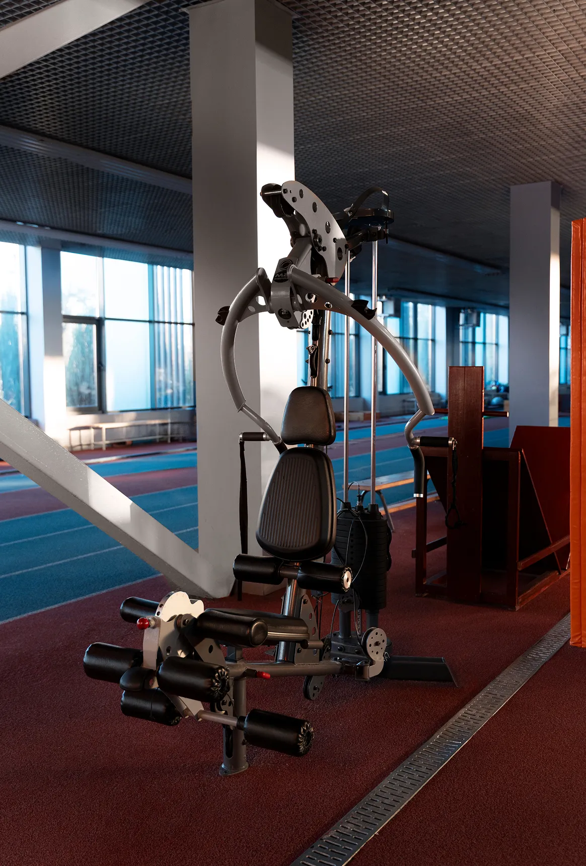 Multi-function gym machine with padded seat and handles in an indoor sports facility with large windows and running tracks.