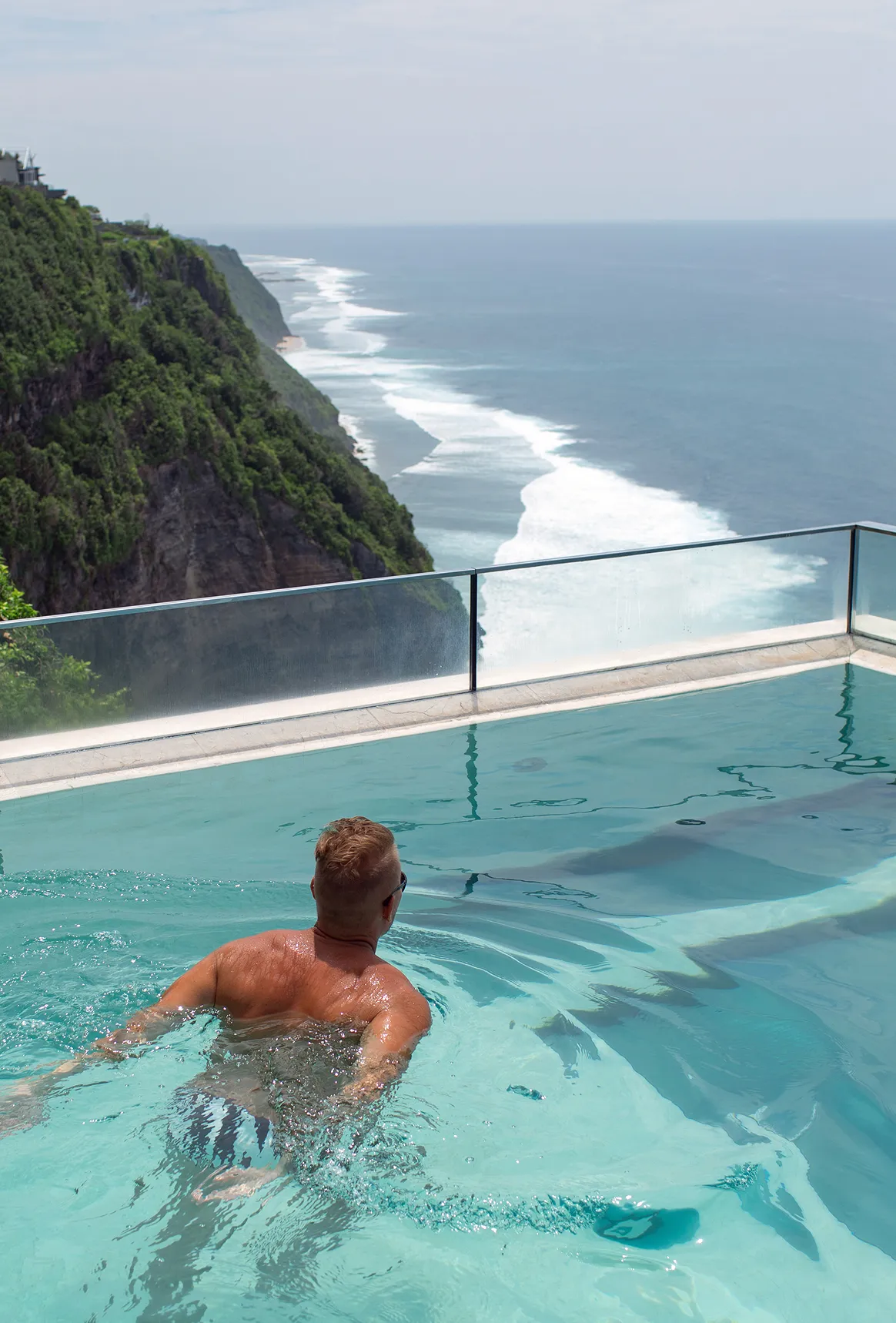 Man swimming in a pool overlooking a cliffside with ocean waves in the background.