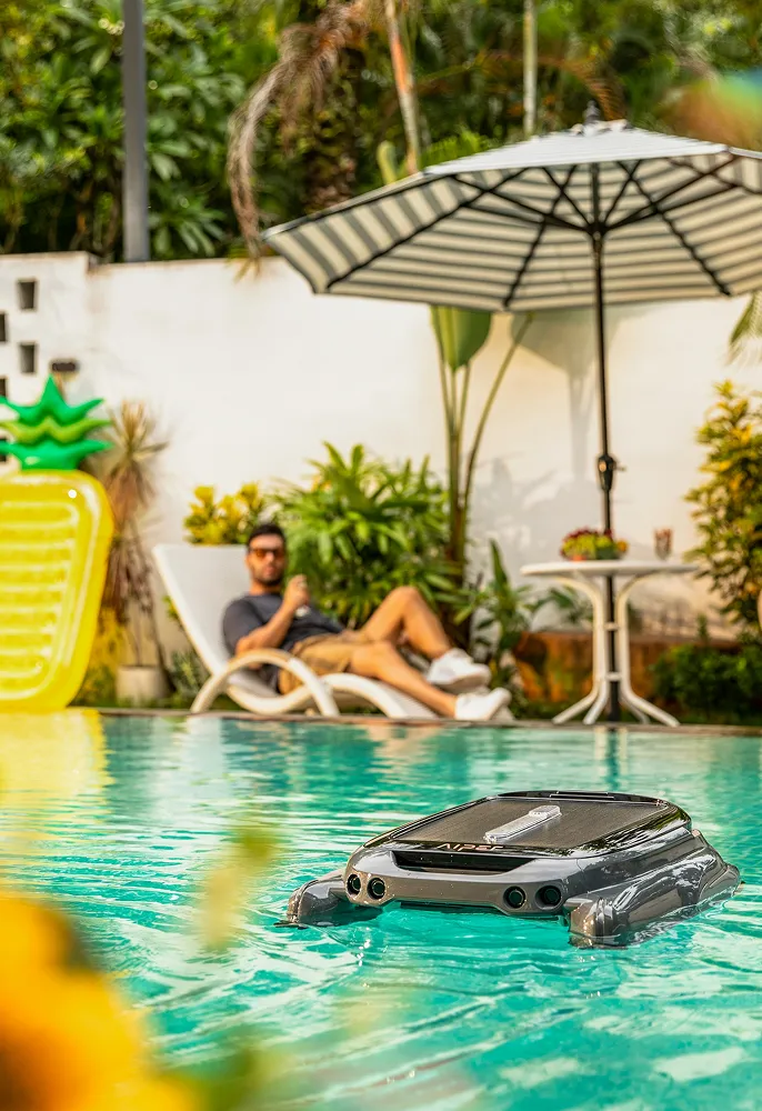 Robotic pool cleaner floating on clear blue water with a man relaxing on a lounge chair under a large umbrella in the background.