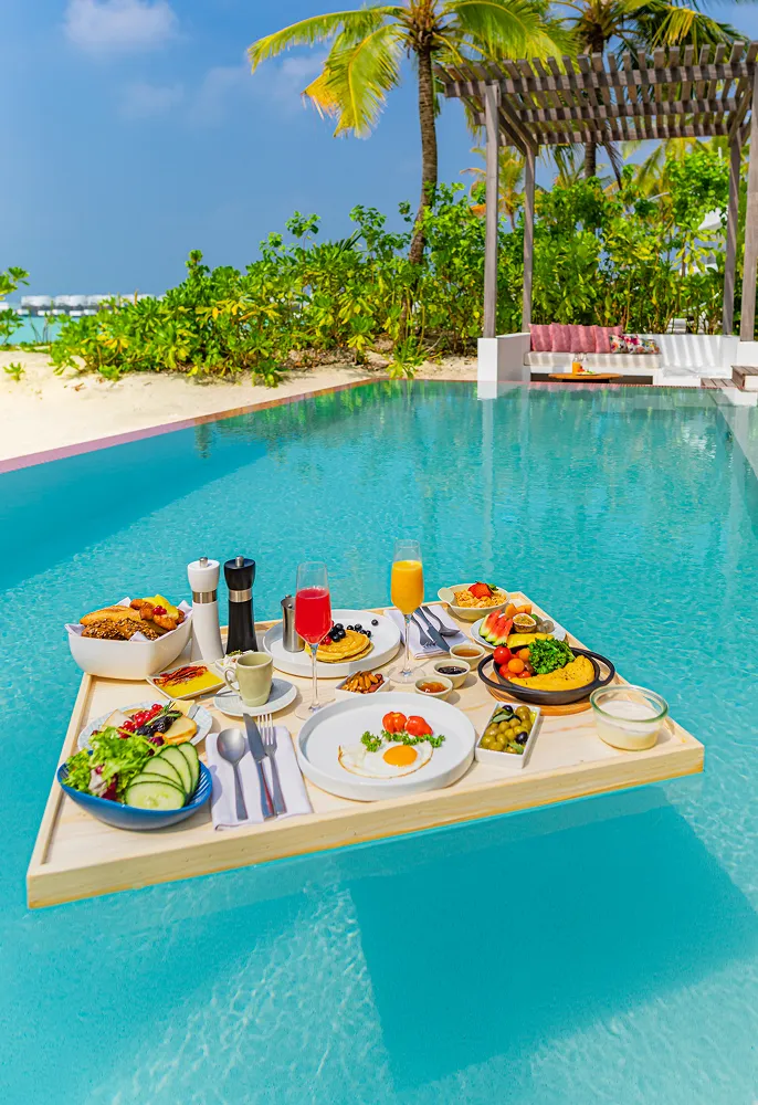 Floating breakfast tray with assorted dishes and drinks on a pool with palm trees and beach in the background.
