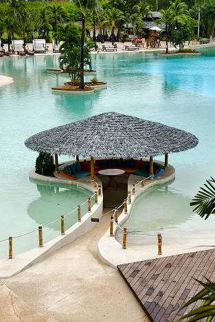 Thatched-roof poolside bar surrounded by water with seating inside and palm trees in the background.