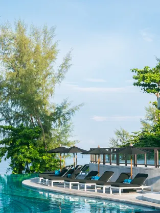 Curved outdoor poolside with lounge chairs and umbrellas surrounded by trees under a clear sky.