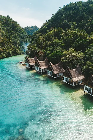 A row of thatched-roof huts on stilts along a clear turquoise river surrounded by dense green forested hills.