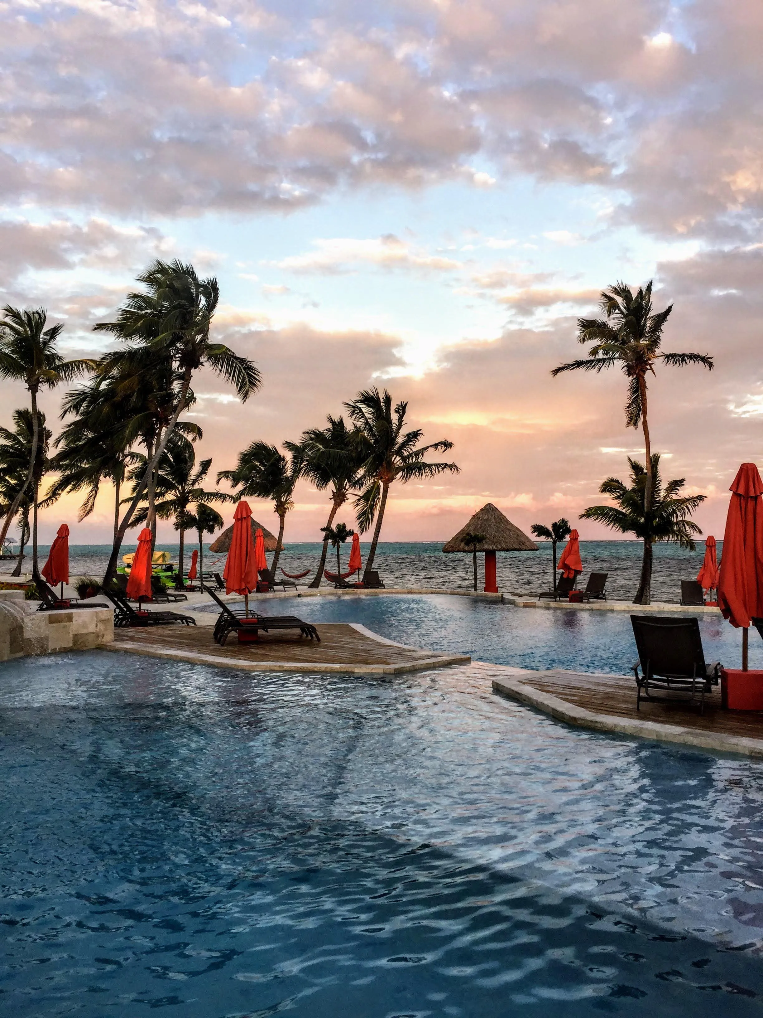 Resort pool with palm trees and red umbrellas at sunset by the ocean.