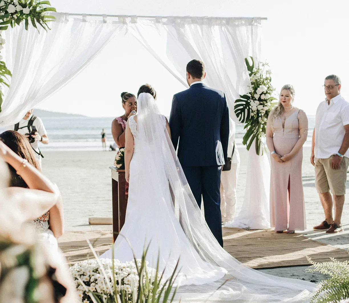 Bride and groom standing under a white arch with flowers during a beach wedding ceremony.