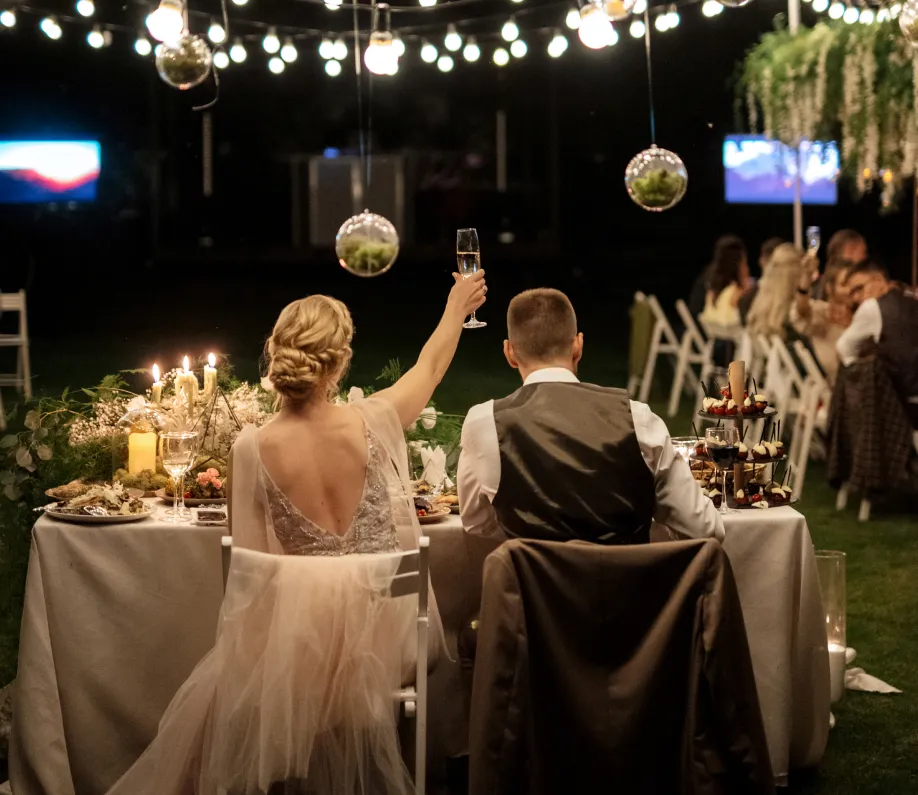 Bride raising a champagne glass at a decorated outdoor wedding reception with guests seated in the background.