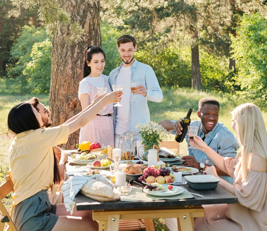 Group of six friends enjoying an outdoor meal in a sunny park, raising glasses for a toast around a table with food and drinks.