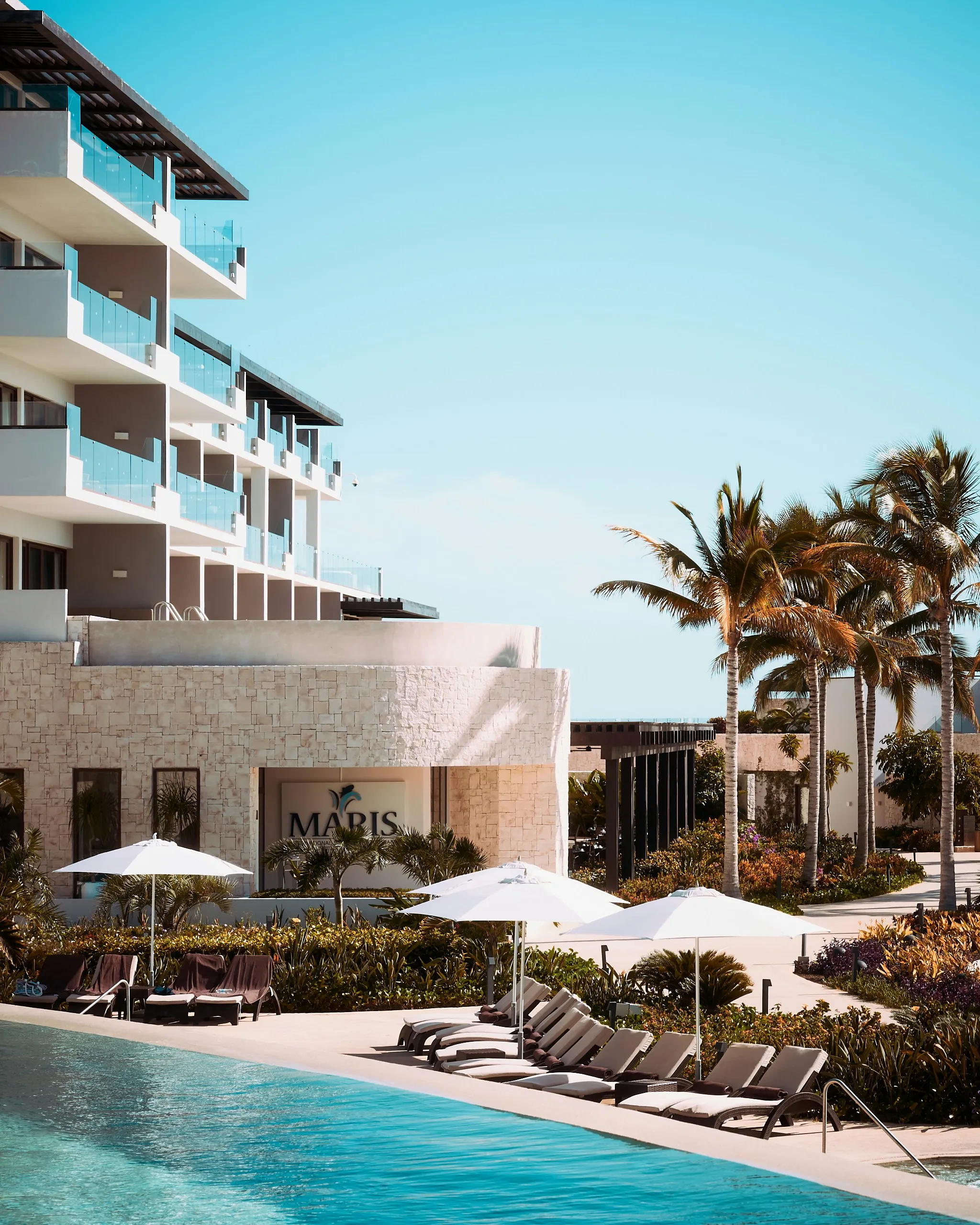 Poolside lounge chairs with white umbrellas next to a modern hotel building and palm trees under a clear blue sky.
