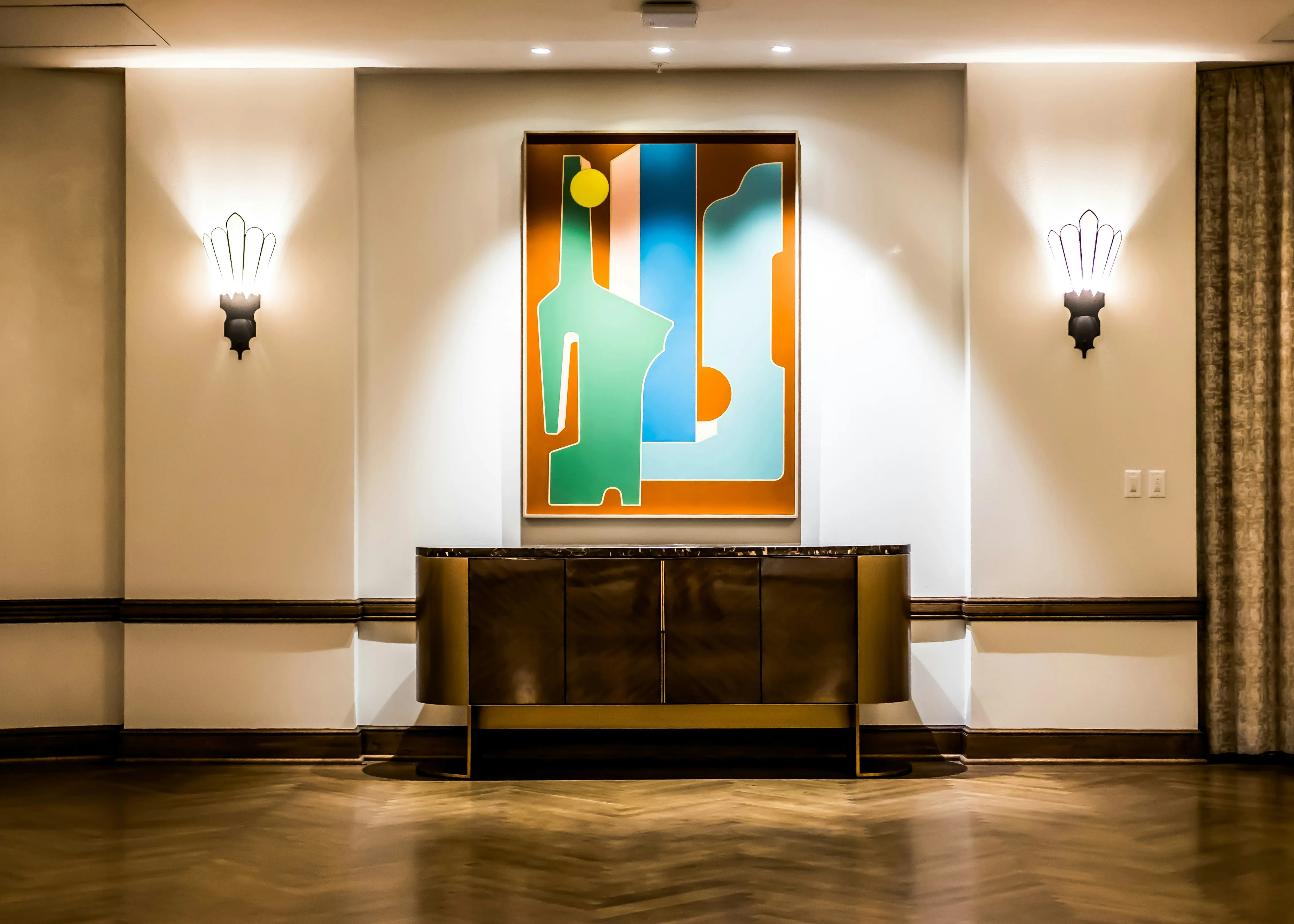 Modern interior with a wooden sideboard beneath an abstract colorful painting, flanked by two wall sconces.