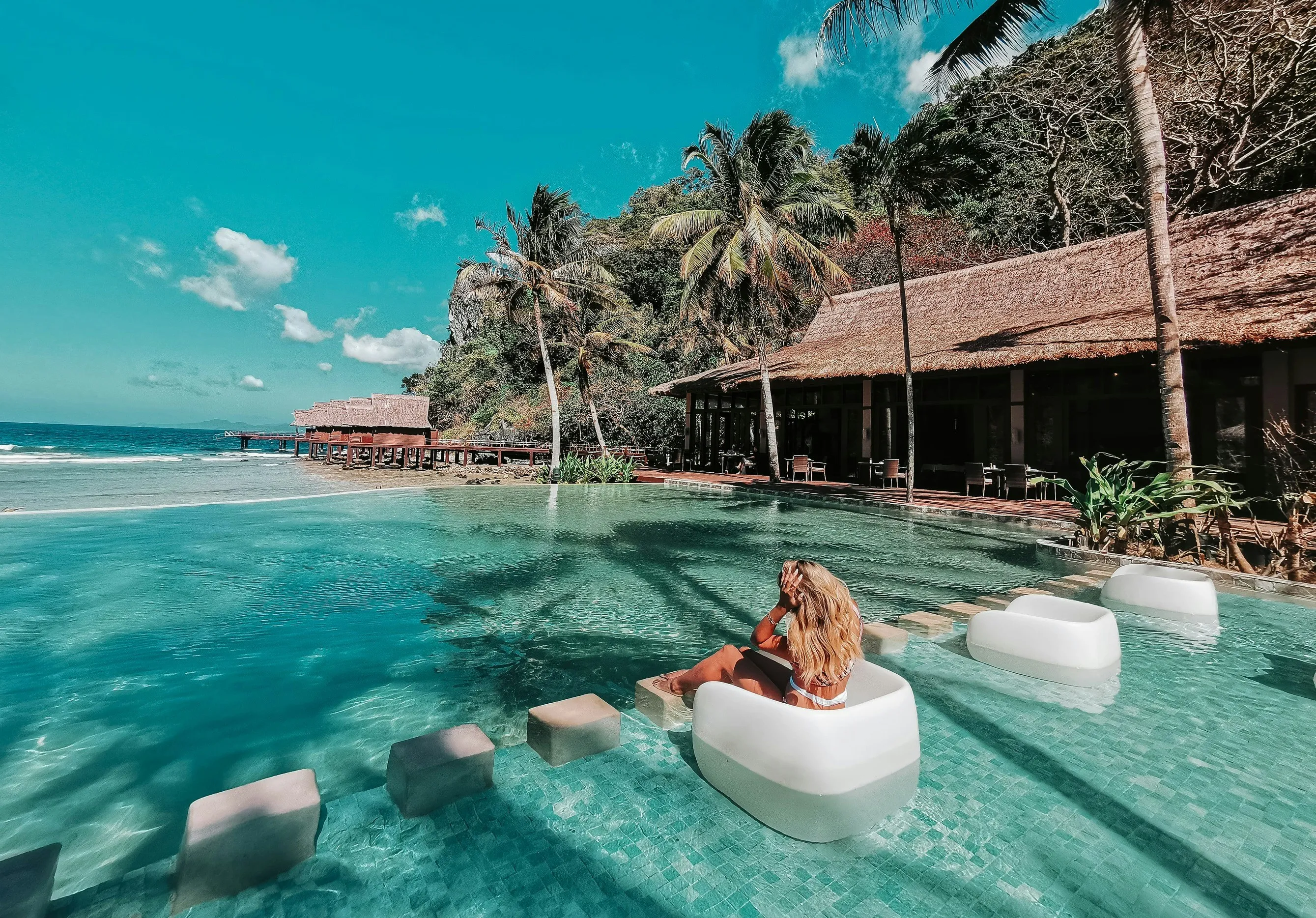 Woman in white swimsuit sitting on a white poolside chair in an infinity pool with palm trees and thatched buildings in the background.