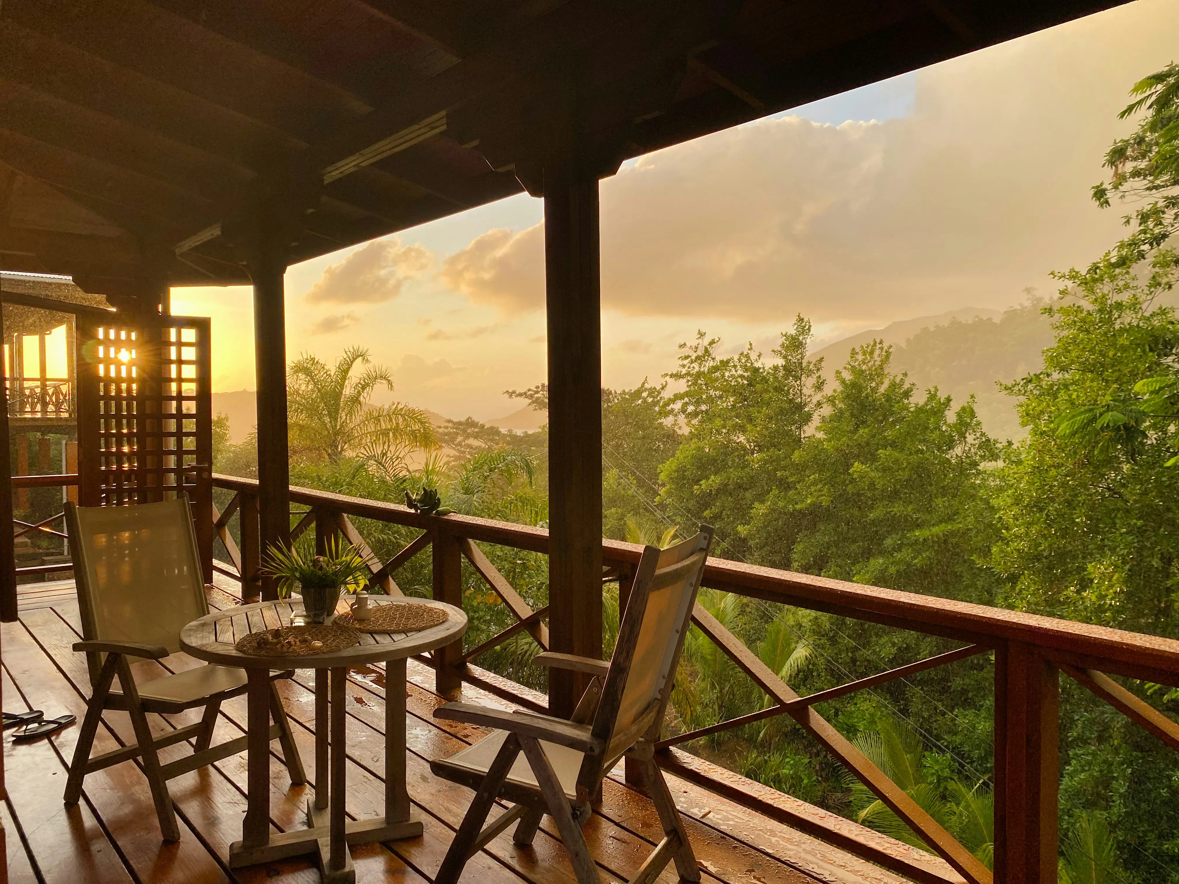 Wooden balcony with two chairs and a round table overlooking lush green trees and distant mountains at sunset.
