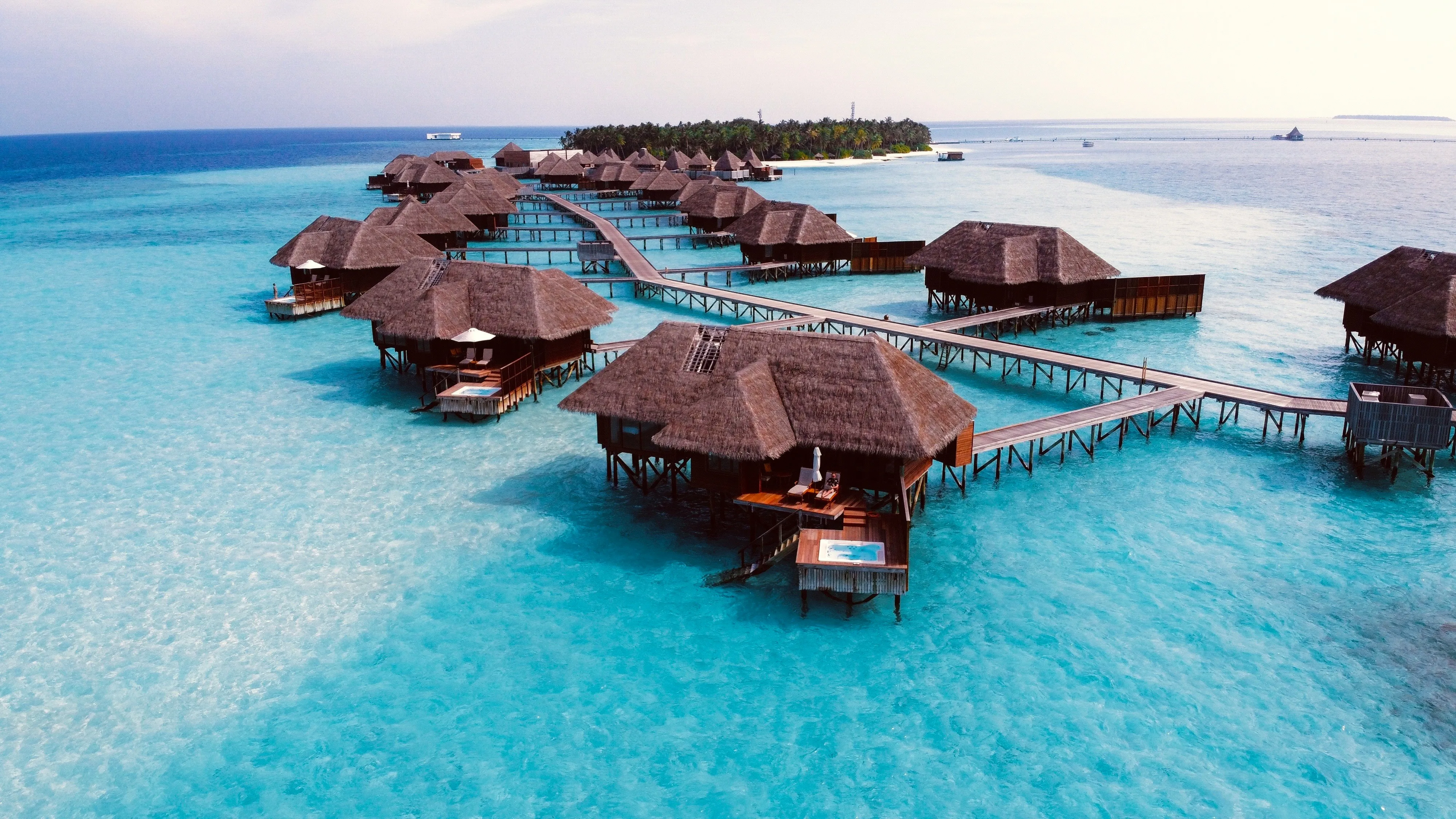 Overwater bungalows connected by wooden walkways over clear turquoise ocean water with a small island in the distance.