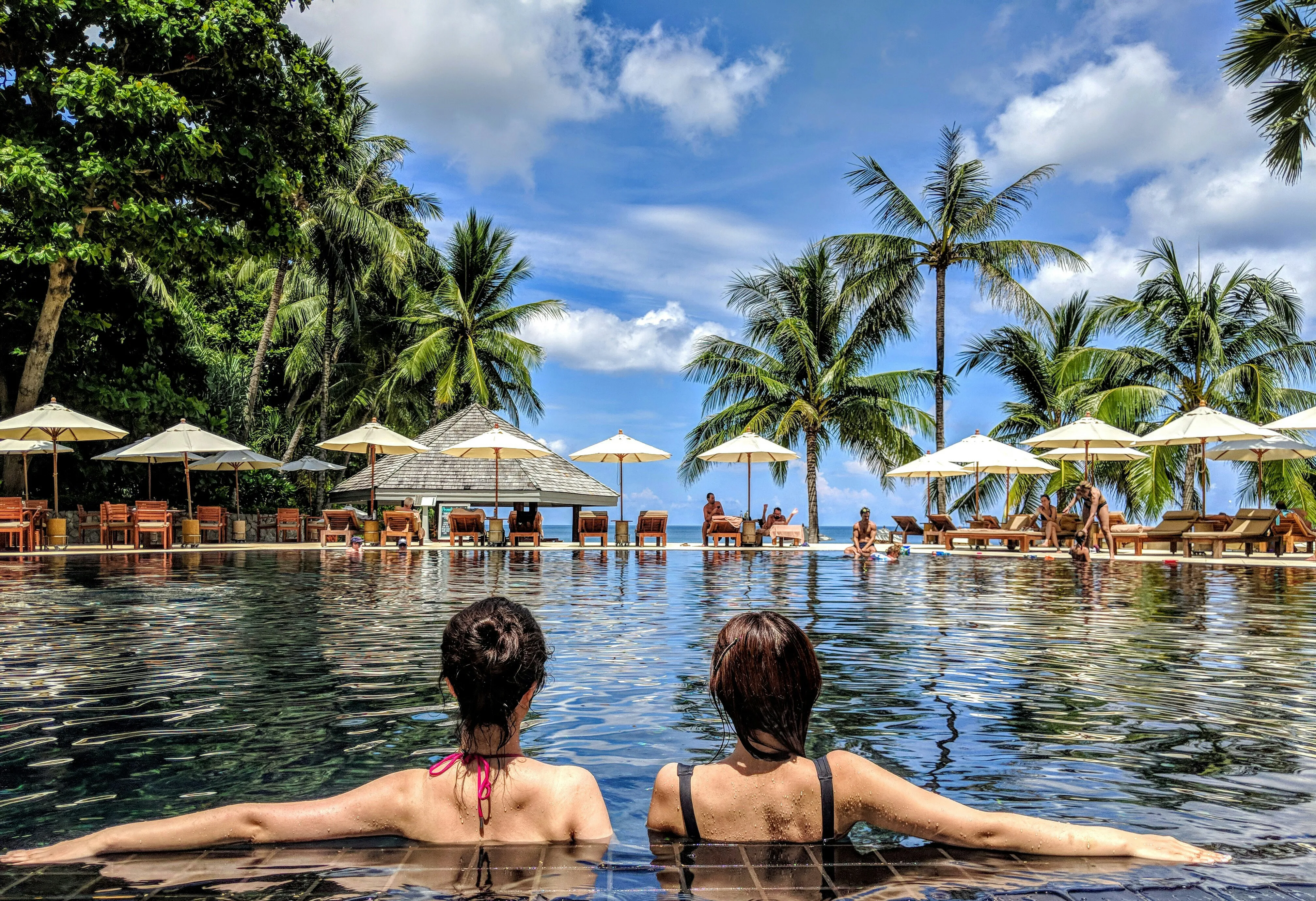 Two women relaxing in a resort swimming pool with umbrellas, palm trees, and lounge chairs in the background under a partly cloudy sky.