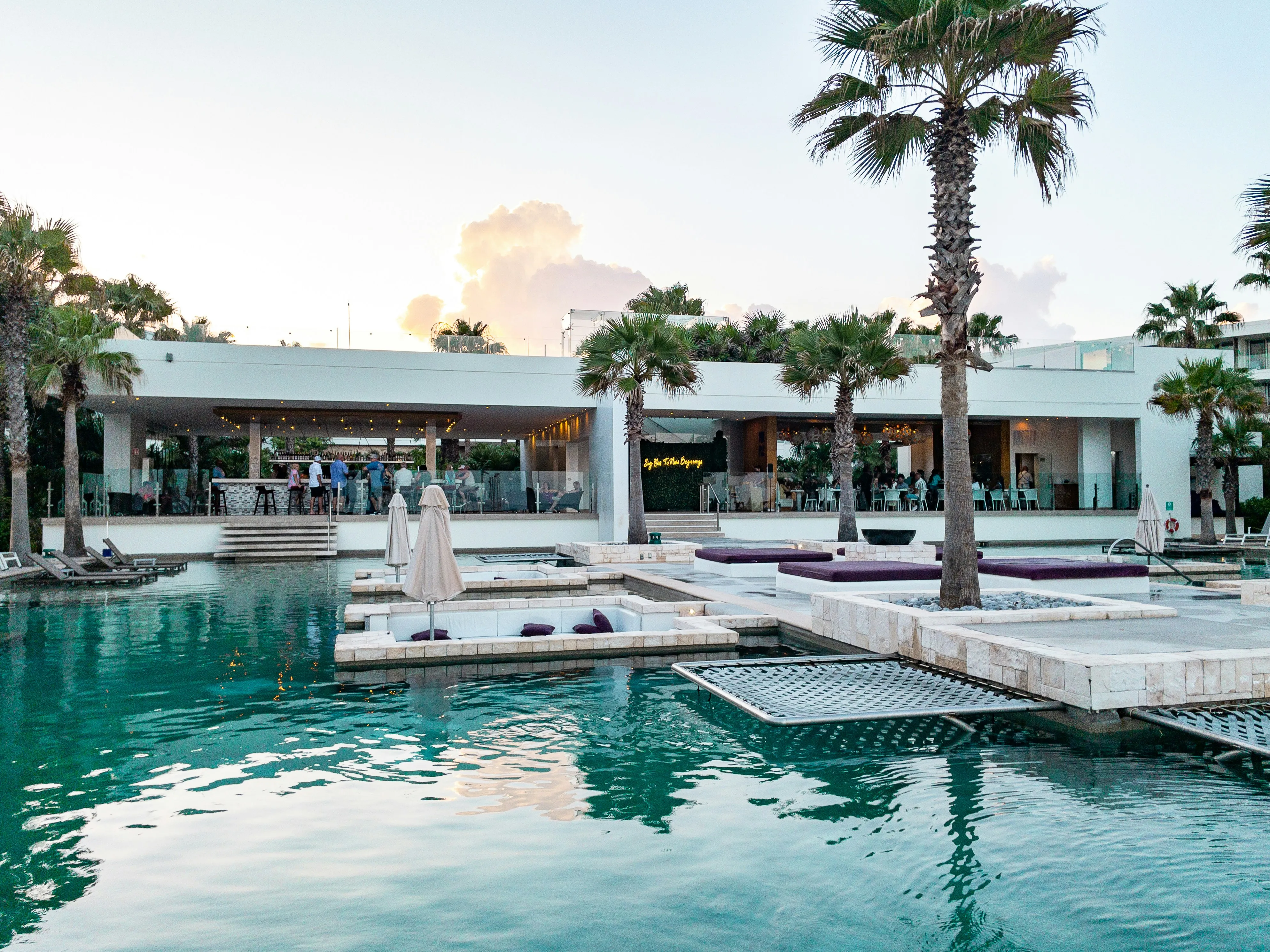 Modern outdoor pool area with sunken seating, palm trees, and people dining in a nearby open-air restaurant at sunset.