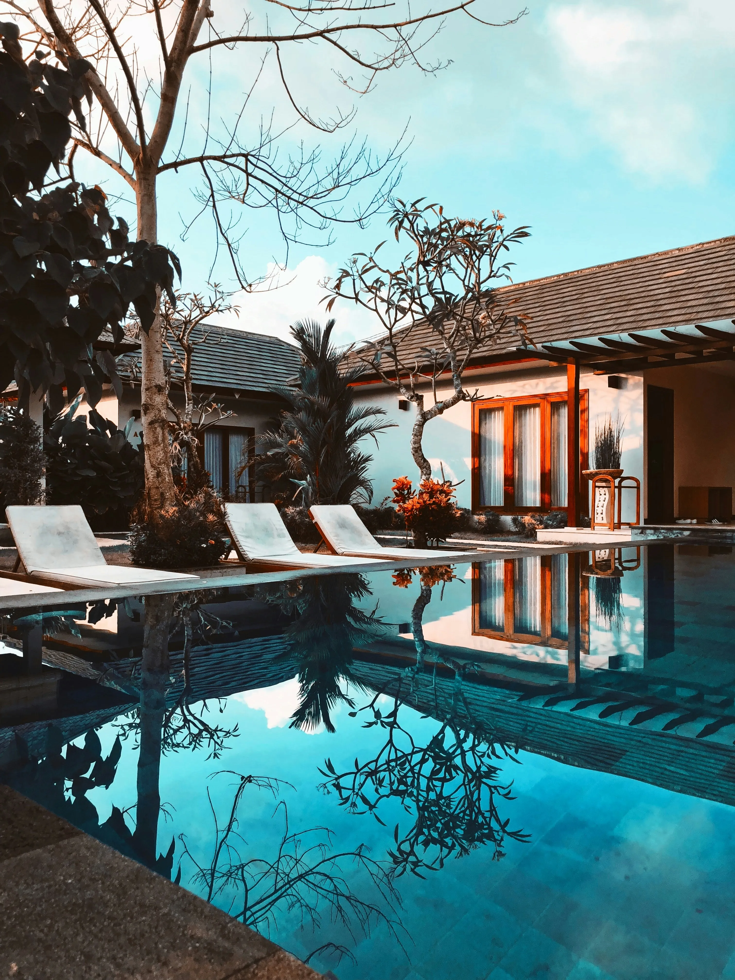 Poolside view of a modern house with lounge chairs, trees, and clear blue water reflecting the sky and surrounding foliage.