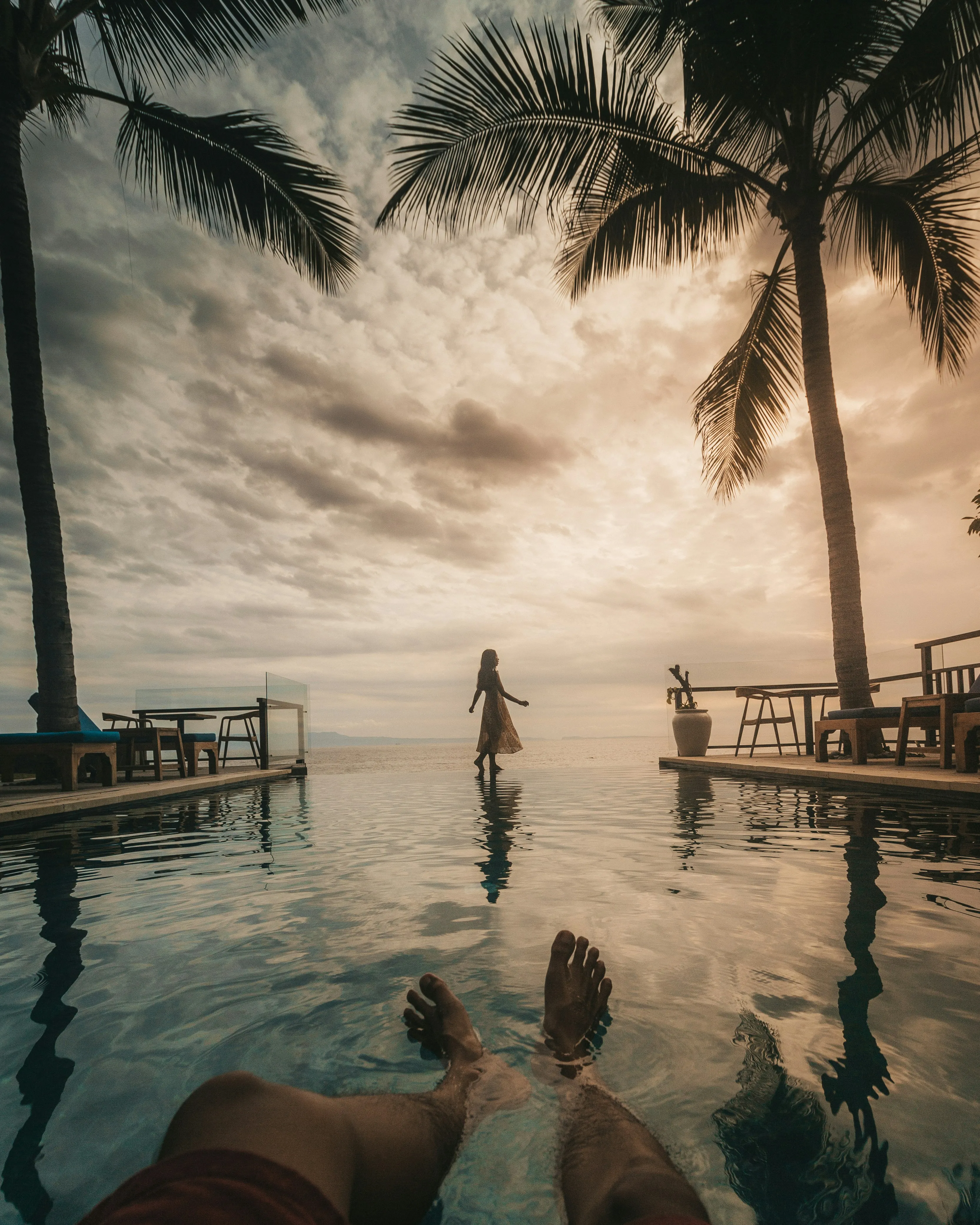 View from a pool with legs in water, palm trees on sides, and a woman walking near an edge with a sunset sky.