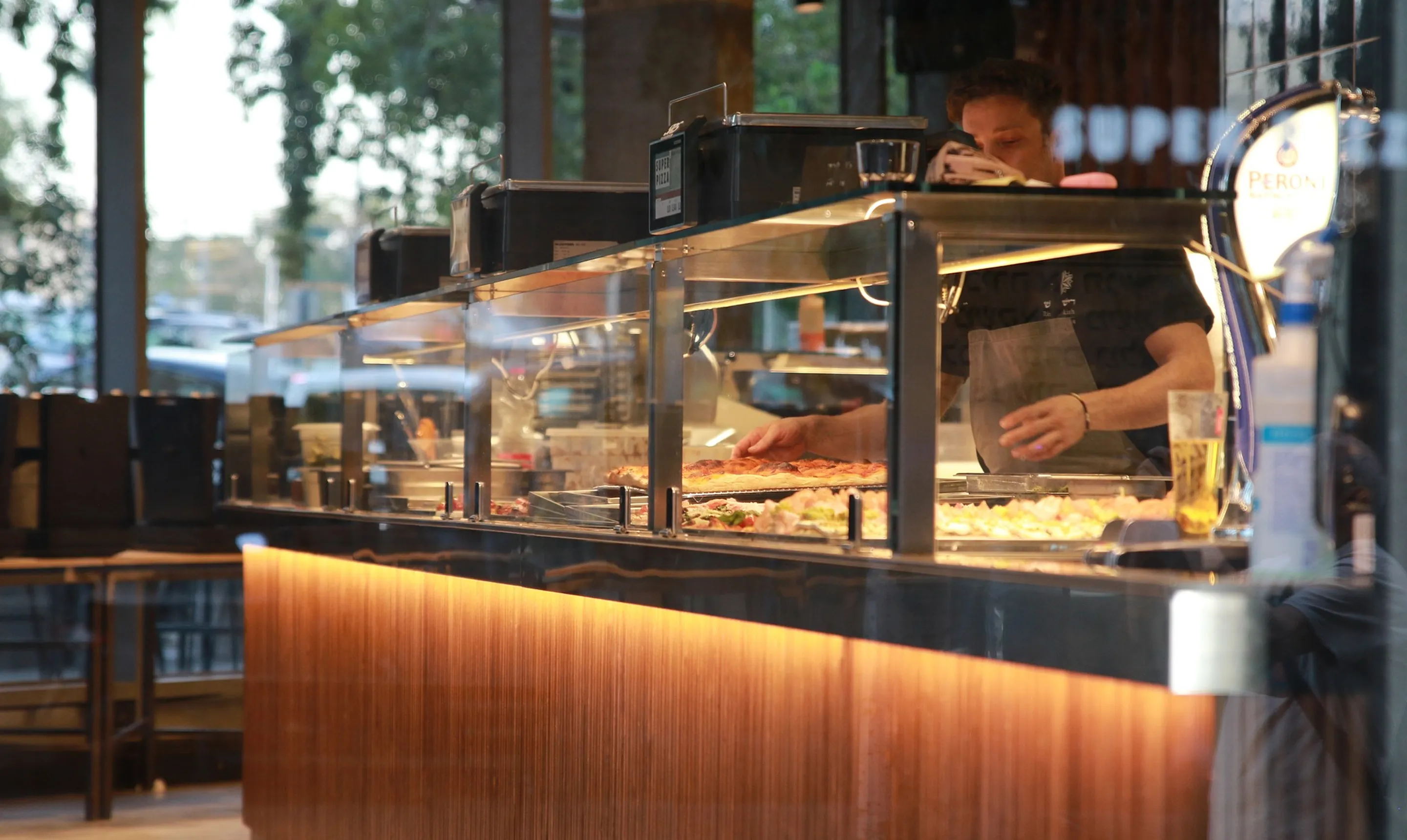 Person behind glass counter serving slices of pizza in a restaurant with warm interior lighting.