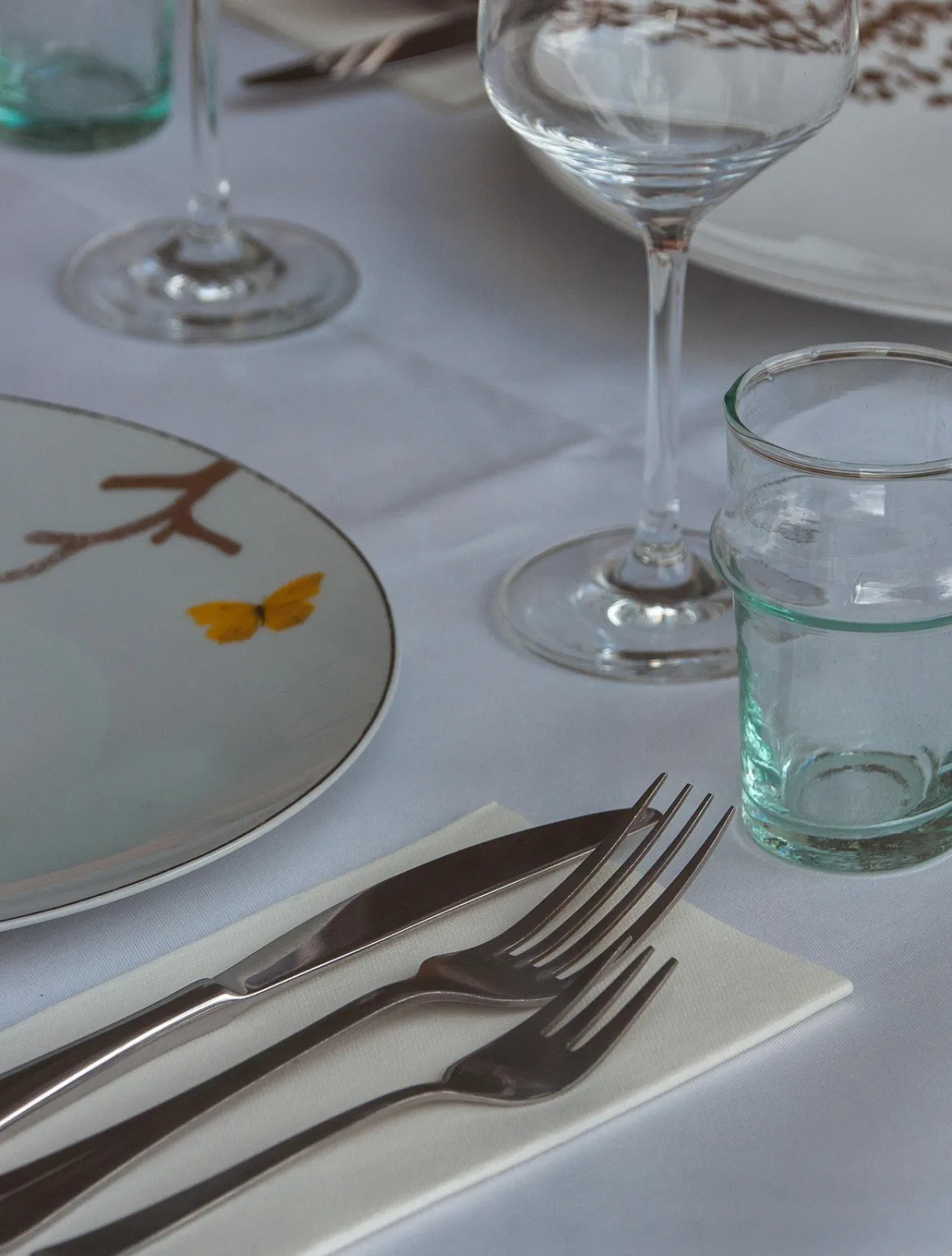 Close-up of a table setting with two forks, a knife on a folded white napkin, a plate with a yellow butterfly design, a wine glass, and a water glass on a white tablecloth.